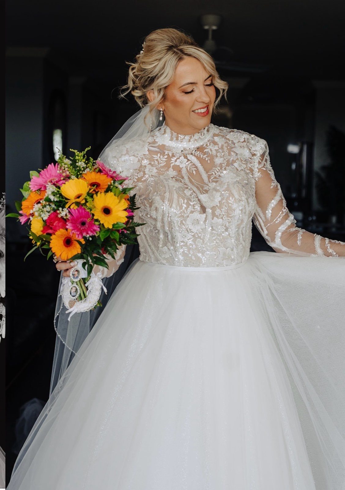 A bride looks down and smiles in a gown with a high-neck illusion bodice heavily beaded with floral lace, long sheer sleeves and a full sparkling tulle skirt. A veil falls behind her and she holds a vivid bouquet of pink and orange gerberas and sunflowers against a dark interior.