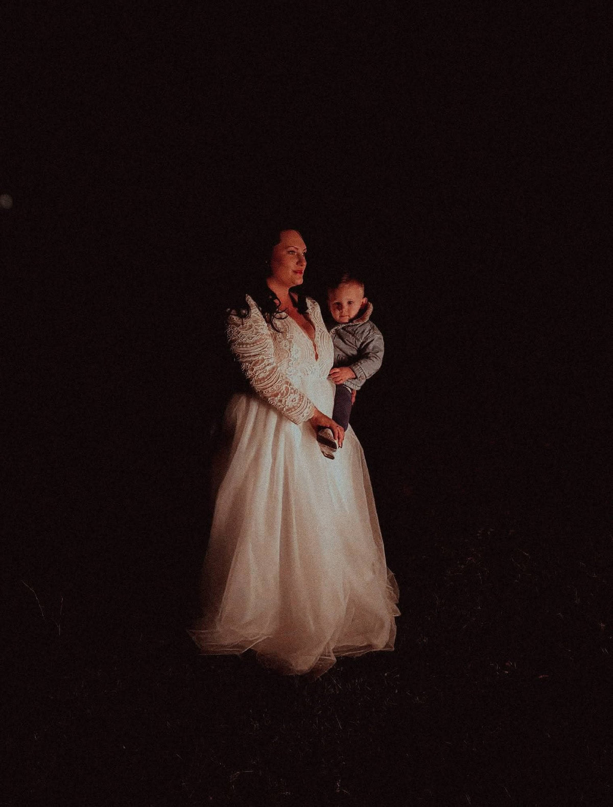A bride stands lit against a black night background, holding a toddler on her hip. Her gown has long lace sleeves and a deep V neckline over a fitted lace bodice, falling into a full, softly gathered tulle skirt.