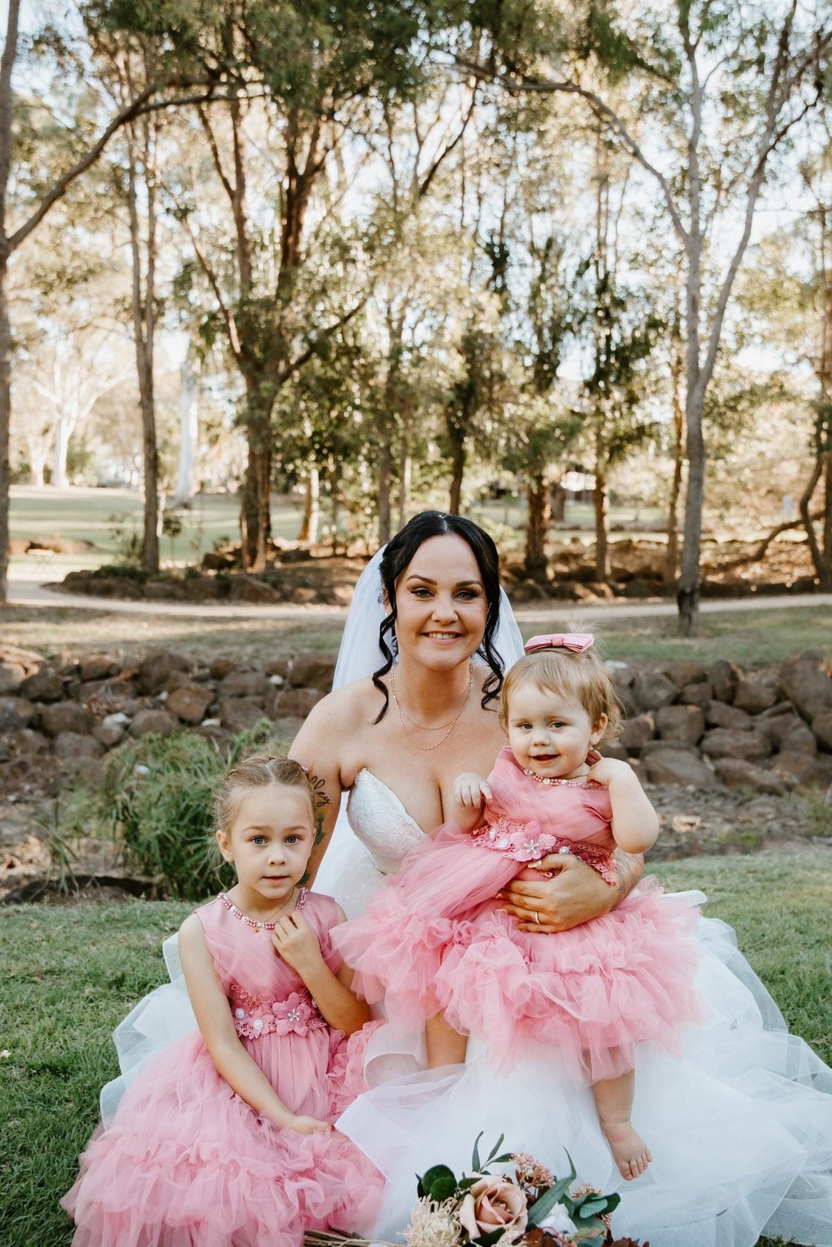 A bride kneels on the grass in a strapless white gown with a full tulle skirt and veil, her arm around two small flower girls in ruffled pink tulle dresses. Dry bushland and gum trees fill the background, with a stone retaining wall behind them and her bouquet resting on the grass.