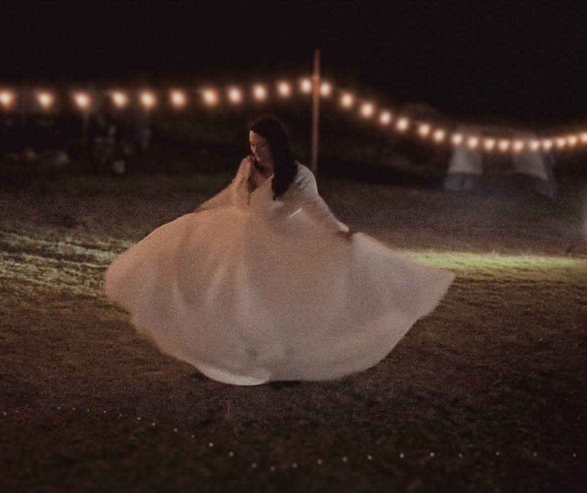 A bride twirls on grass at night beneath a strand of festoon lights, the full skirt of her wedding gown flared out wide around her. The photograph is warm and grainy, with the lights and a marquee glowing softly in the dark behind her.
