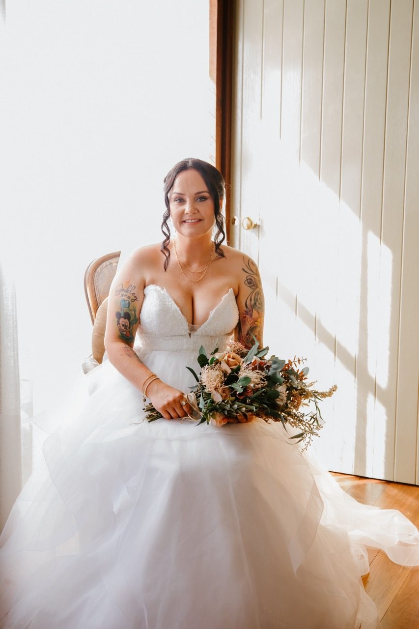 A bride sits in soft window light in a strapless gown with a fitted lace bodice and a layered tulle skirt falling around the chair. She holds a native bouquet of roses, banksia, eucalyptus and dried grasses, with sunlight striping the panelled wall behind her.