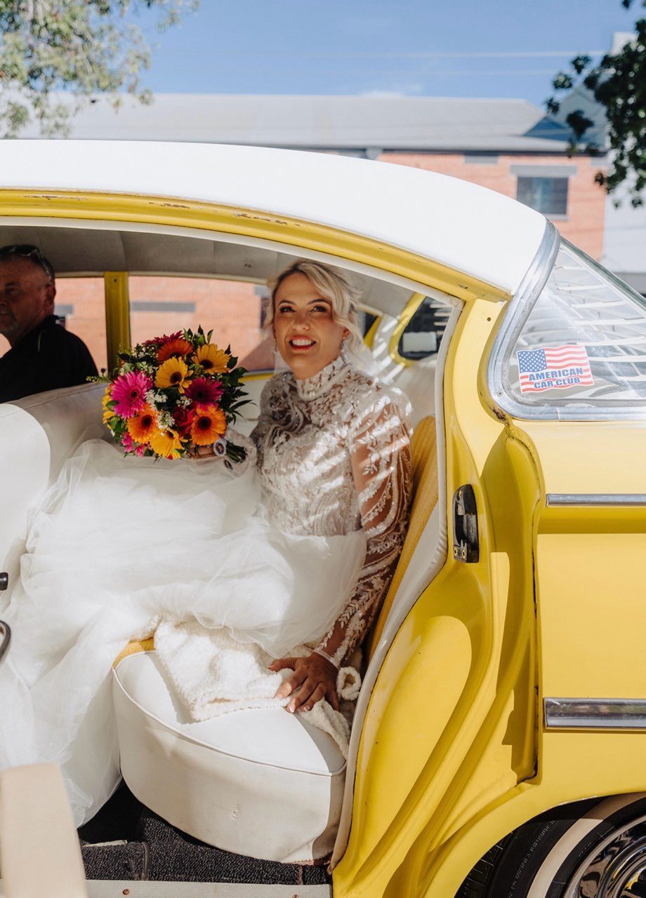 A bride sits in the open door of a yellow and white vintage Chevrolet, smiling up out of frame. Her gown has a beaded high-neck bodice with long sheer lace sleeves, and she holds a bright bouquet of gerberas and sunflowers in pink, orange and yellow.