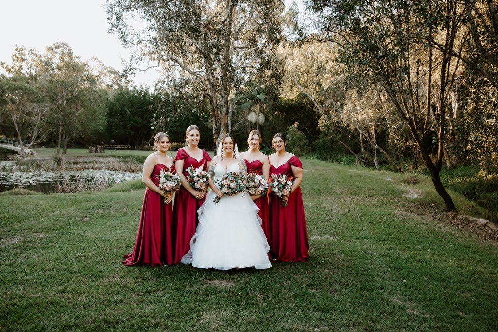 A bride in a tiered tulle ballgown stands on a grassy bank beside a lily pond at golden hour, flanked by four bridesmaids in deep red satin gowns with off-shoulder and sweetheart necklines. All five hold soft bouquets of blush roses and eucalyptus, with gum trees rising behind them.