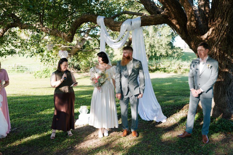 An outdoor ceremony beneath a large spreading tree hung with white draping. The bride stands in her ivory satin puff-sleeve gown holding a native bouquet, the groom beside her in a sage suit, with the celebrant speaking into a microphone on one side and a groomsman on the other, on a sunlit lawn.