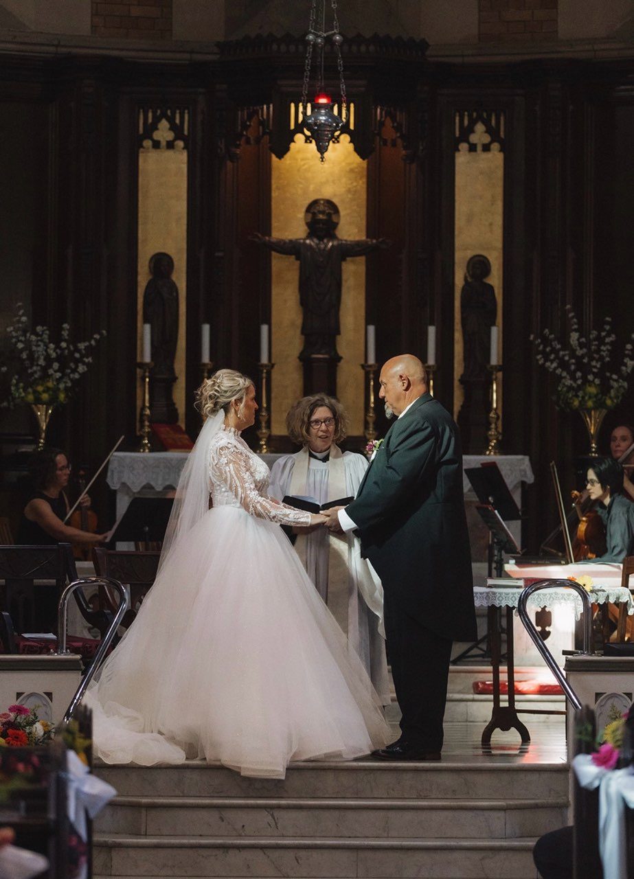 A church ceremony at a dark timber altar with a gilded gold panel behind. The bride wears a long-sleeved lace bodice and a full blush tulle ballgown skirt, holding hands with her groom in a deep green jacket while the celebrant reads between them and string players sit either side.