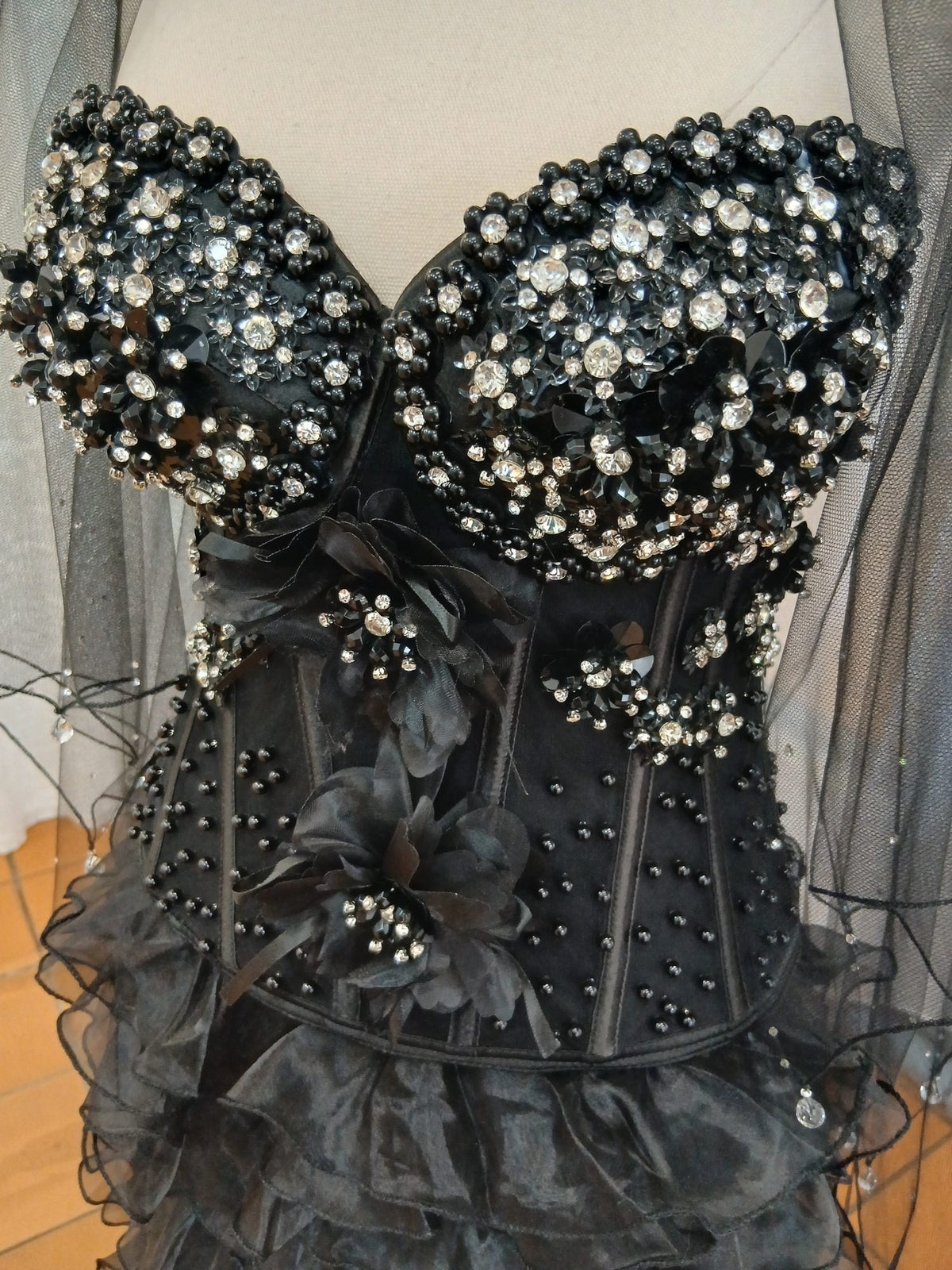 Close-up of the bodice of a strapless black formal gown, densely hand-beaded across the bust with jet pearls and clear crystals, with black organza flowers and scattered beading over the boned waist above a ruffled organza skirt.