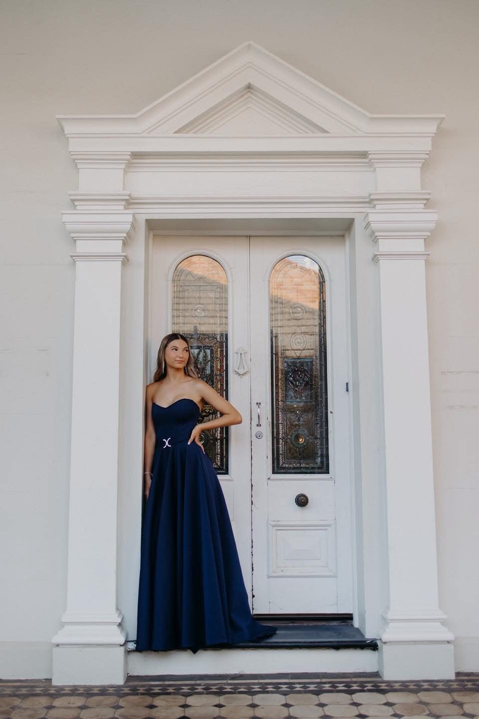 A young woman stands in the recessed white doorway of a heritage building, wearing a strapless navy formal gown with a sweetheart neckline, a full skirt and a short train pooling on the tessellated tiles. She rests one hand on her hip and looks off to the side, framed by painted columns and a pair of timber doors with stained-glass panels.