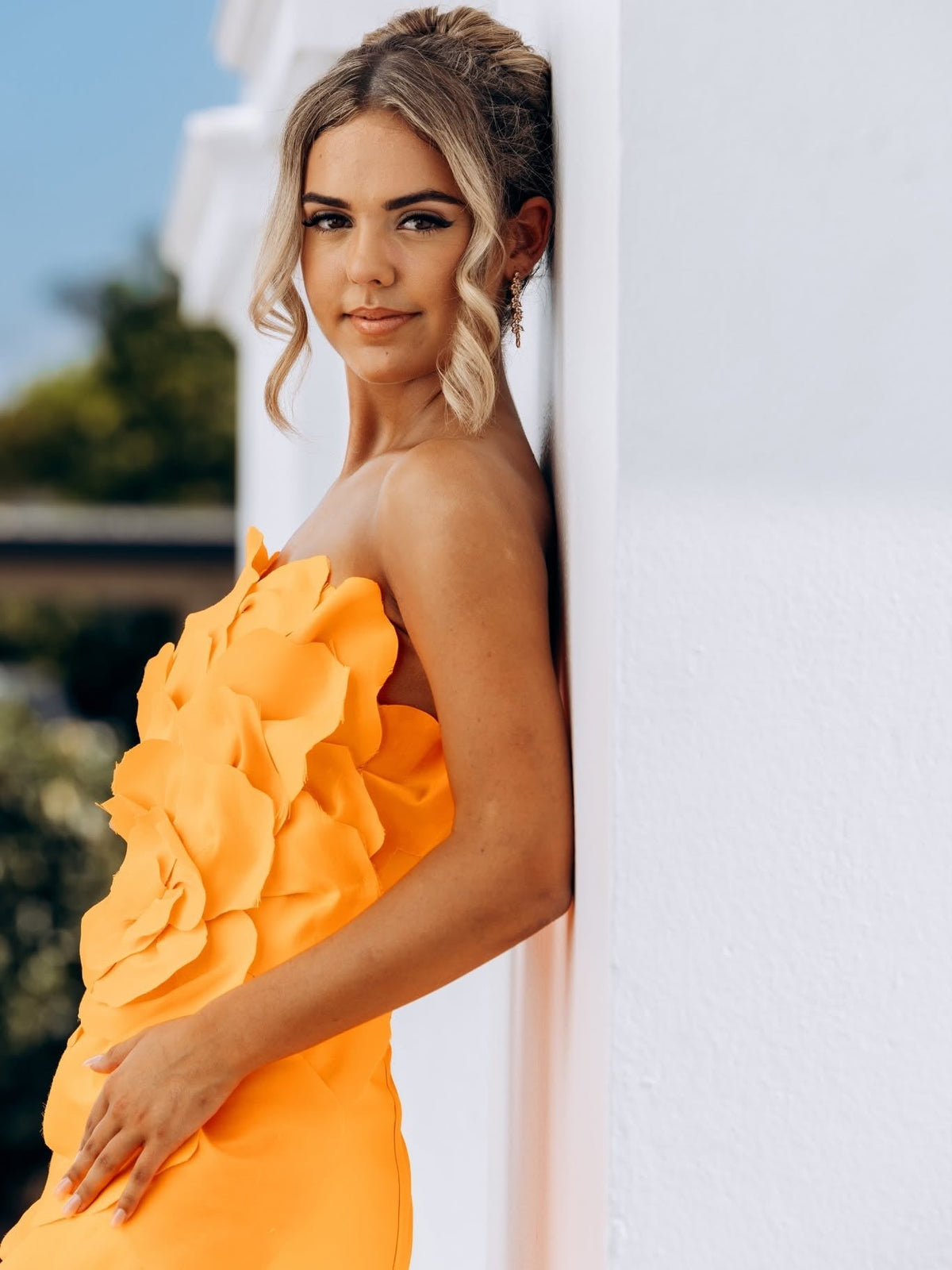 Close portrait of a young woman leaning against a white column in a strapless marigold orange gown, the bodice covered in hand-cut fabric petals