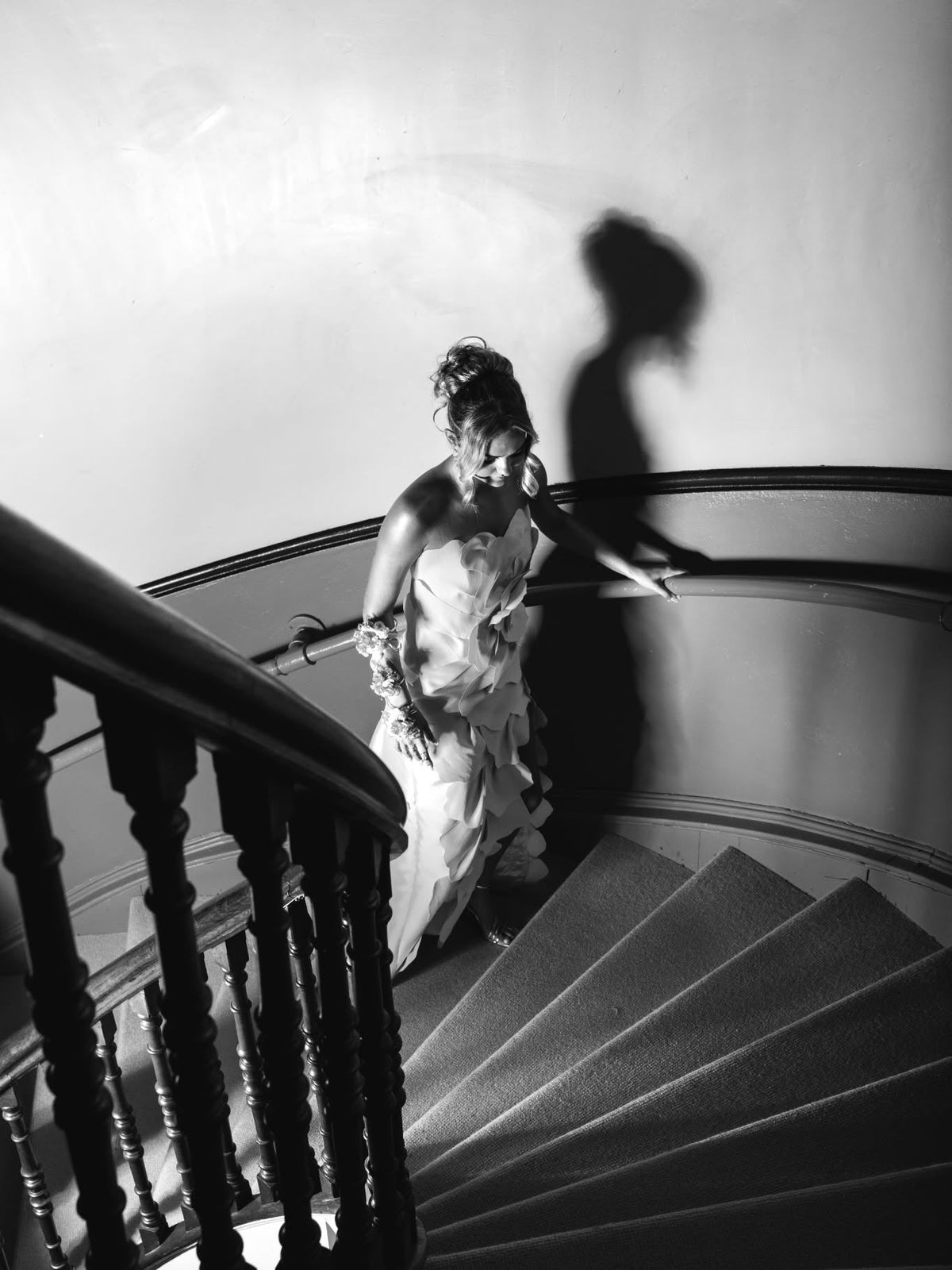 Black and white photograph looking down a curved timber staircase at a young woman in the petal gown, her shadow thrown on the wall behind her