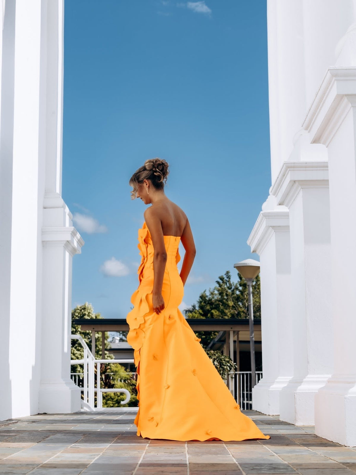 Back view of the marigold orange gown on a tiled terrace between white columns, showing the petal cascade down one side and the fishtail skirt