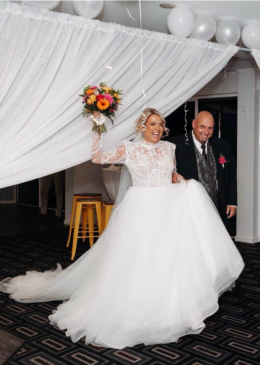 A laughing bride enters her reception under swagged white draping and a cluster of white balloons, one arm raised high with her orange and pink bouquet. She wears a long-sleeved beaded lace bodice and a full tulle ballgown skirt with a train, and is escorted by a man in a black suit and patterned waistcoat.