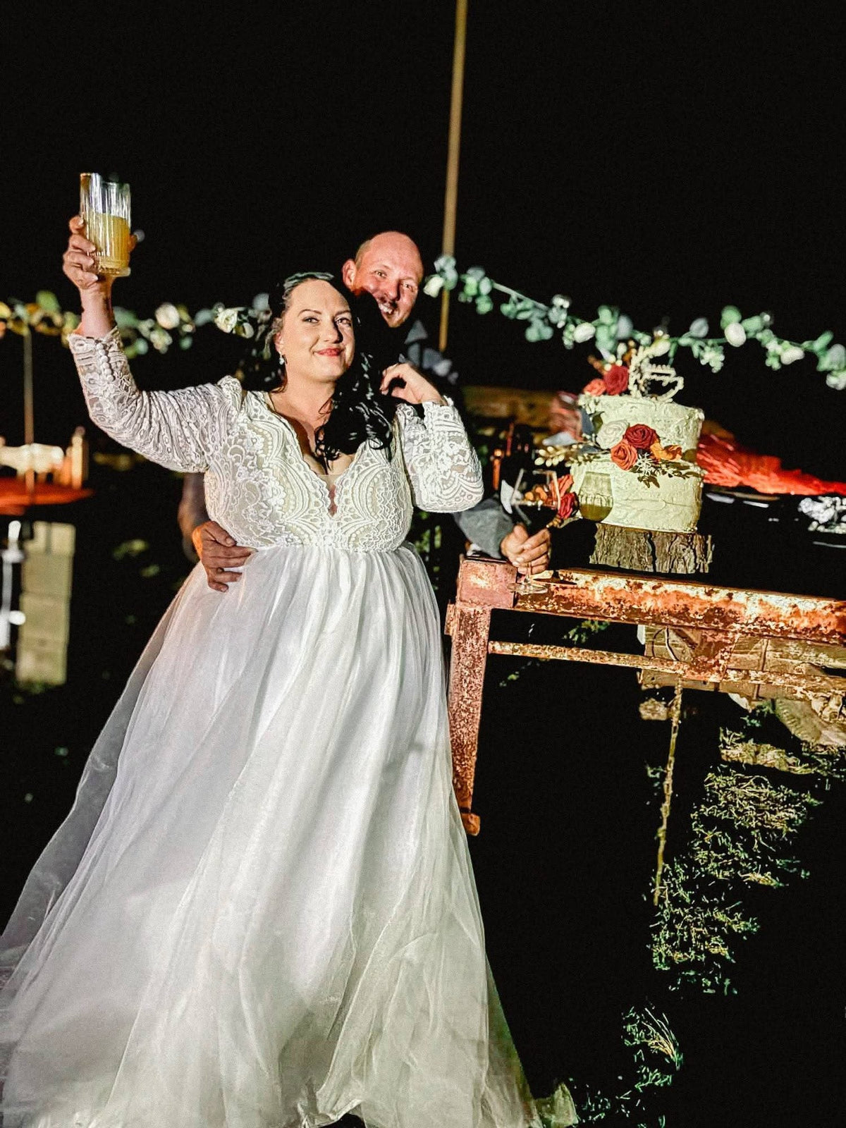 A bride raises a glass in a toast beside her groom at an outdoor reception after dark. She wears a gown with a long-sleeved paisley lace bodice, a V neckline and a soft full tulle skirt, with a tiered wedding cake dressed in roses and a eucalyptus garland on a rustic timber table behind them.