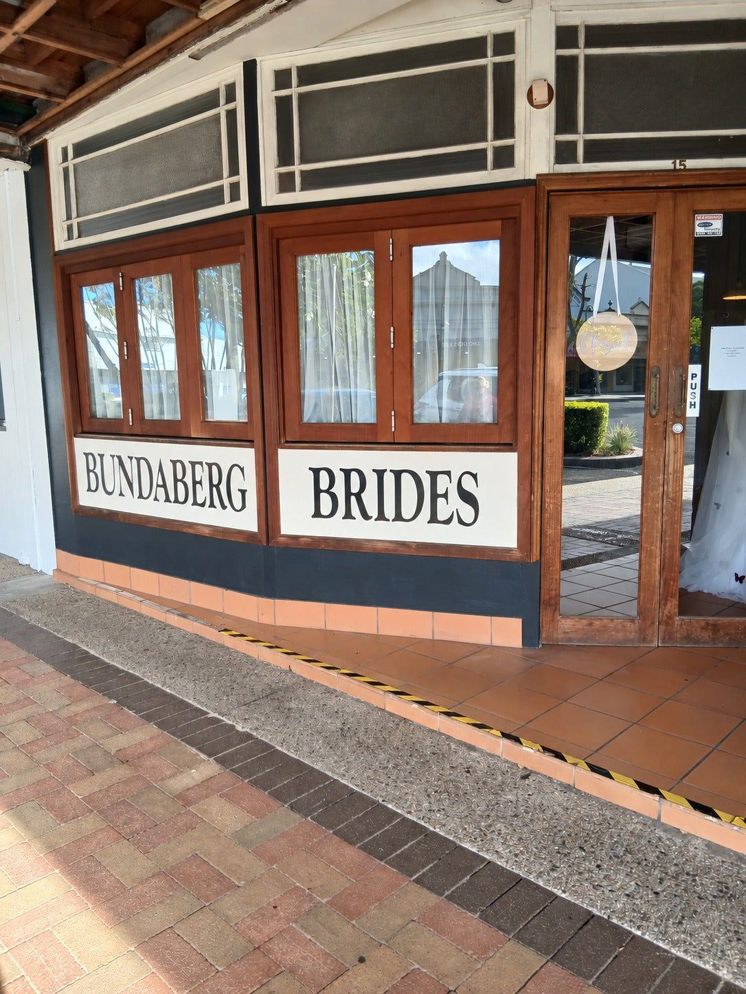 The Bundaberg Brides shopfront on Bourbong Street, with BUNDABERG BRIDES hand-painted in black serif lettering on cream panels beneath timber-framed display windows, and a gown visible through the glass door