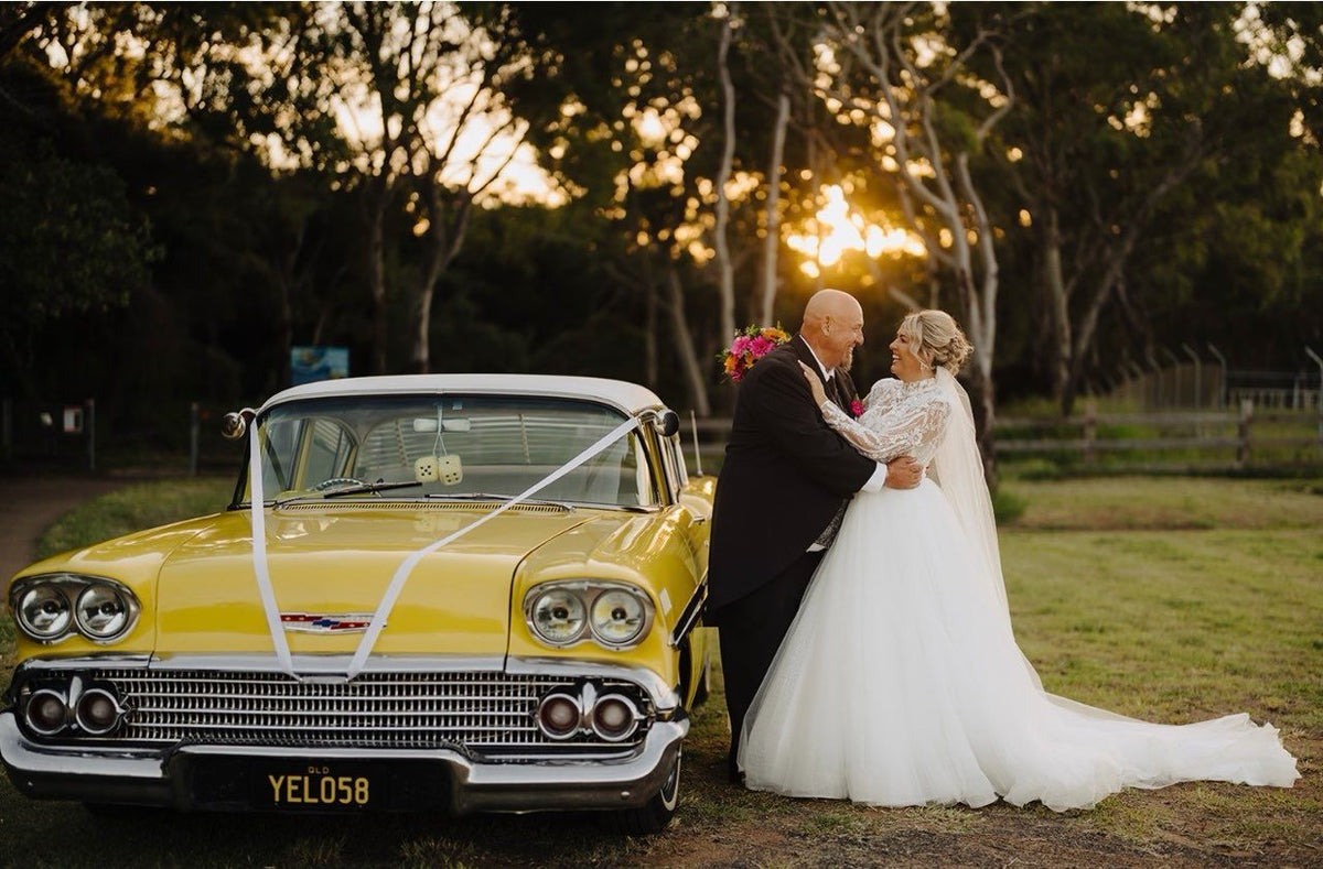 A bride and groom stand embracing beside a yellow vintage Chevrolet dressed with white ribbon, photographed at golden hour with the sun setting through gum trees behind them. Her full tulle skirt and long train spread out across the grass.