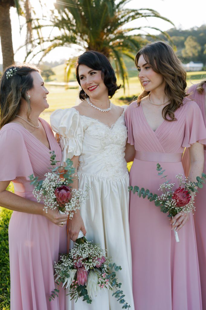 A bride with a dark bob and red lipstick stands between two bridesmaids in late afternoon light, wearing a restored ivory satin gown with full puff sleeves, a beaded sweetheart bodice and a pearl necklace. The bridesmaids wear dusty pink gowns and all three hold bouquets of protea, baby's breath and eucalyptus.