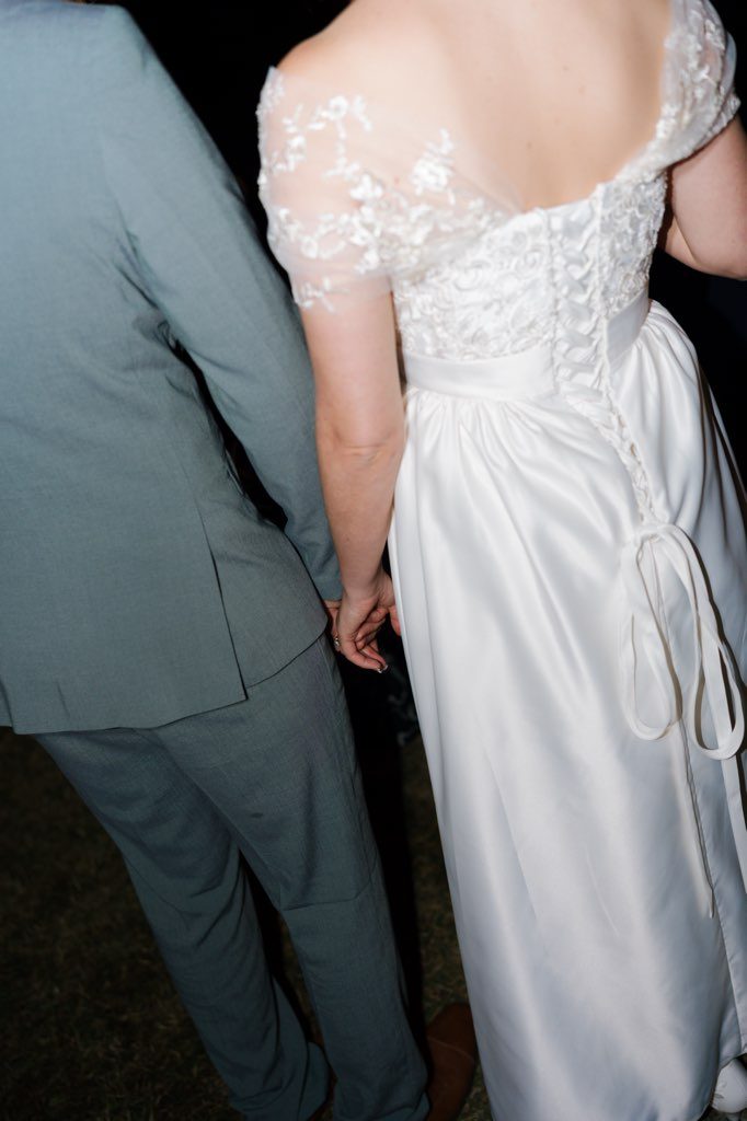A close view of the back of the restored ivory satin gown, showing the fully laced corset back with long satin ties hanging loose, the embroidered lace across the shoulders and the off-shoulder sheer lace sleeves above a smooth gathered satin skirt.