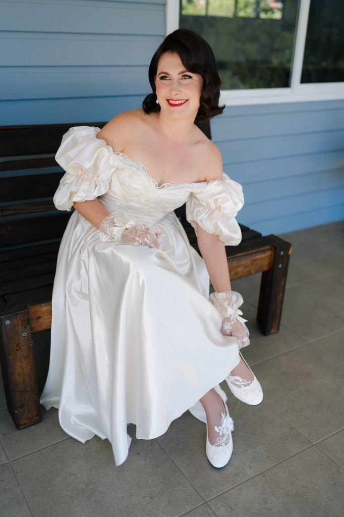 A bride sits on a timber bench against a pale weatherboard wall in the fully restored ivory satin gown, showing the off-shoulder puff sleeves, beaded bodice and sweeping satin skirt. She wears sheer lace gloves and ivory lace shoes, one foot tilted out from under the hem.
