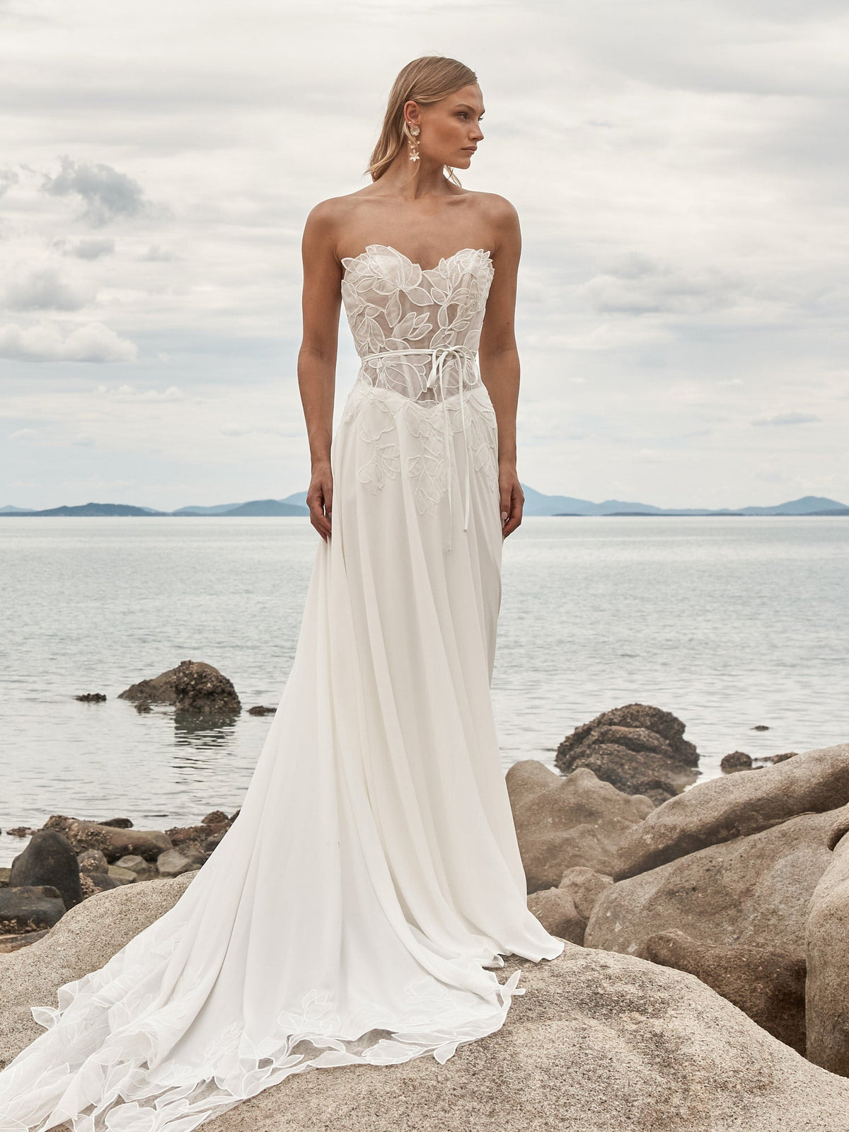 Madi Lane Odyssey gown worn on a rocky shoreline, a strapless sheer lace corset bodice with a tie waist and a flowing chiffon skirt