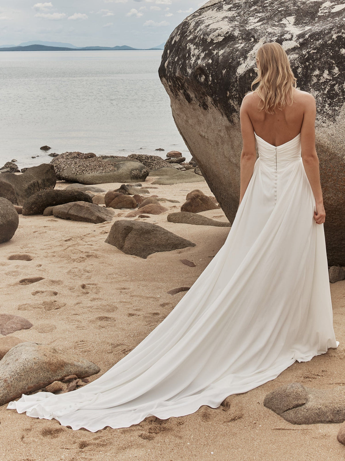 Back view of the Madi Lane Okina gown on a rocky beach, a strapless chiffon gown with a buttoned back and a long sweeping train